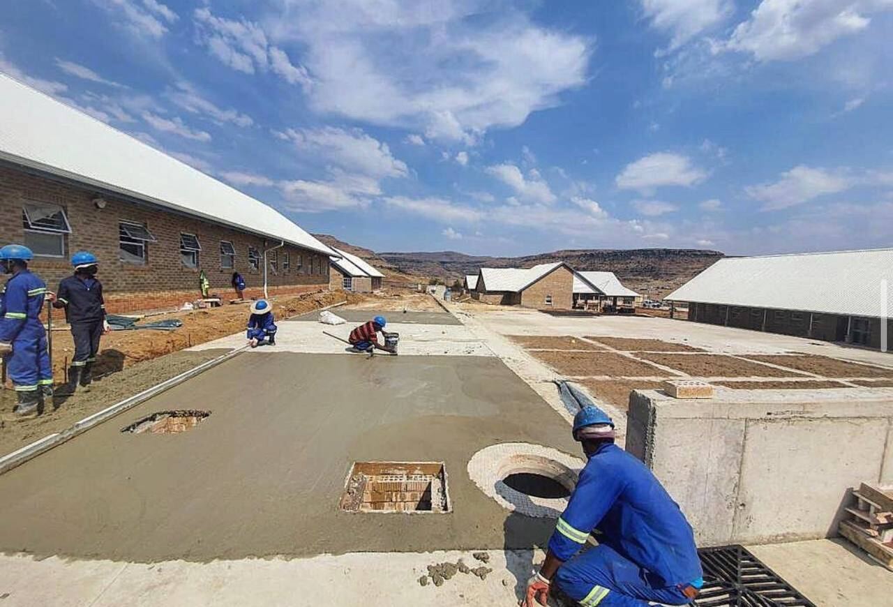 Workers finishing a large concrete bedding slab at an institutional school site with brick buildings in the background